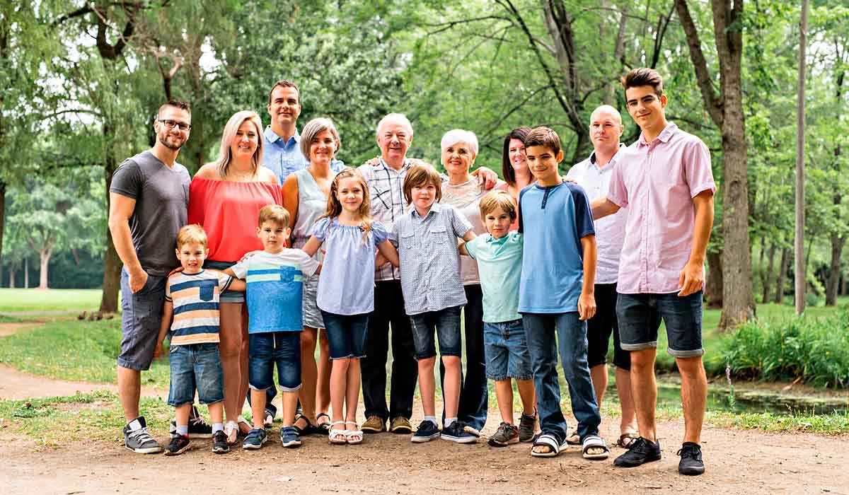 A family stands together outdoors with palm trees and buildings visible in the background. The group consists of two adults and three children, with the mother on the left and the father on the right. They are casually dressed and appear to be in a park or promenade setting.